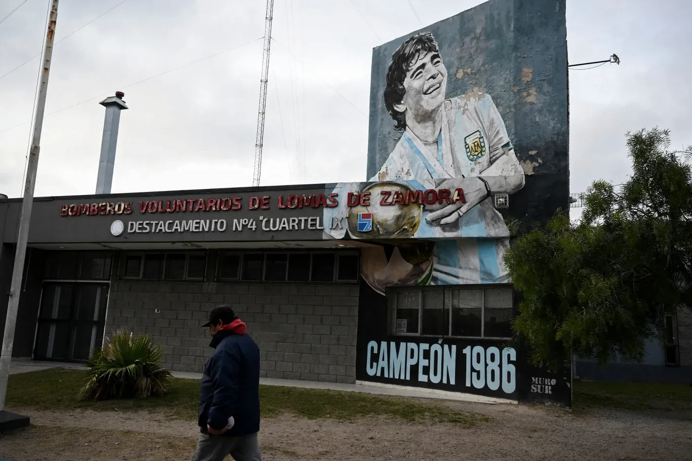 Mural Diego Maradony w stroju Argentyny z napisem "Campeón 1986" na ścianie budynku, obok przechodzi mężczyzna.