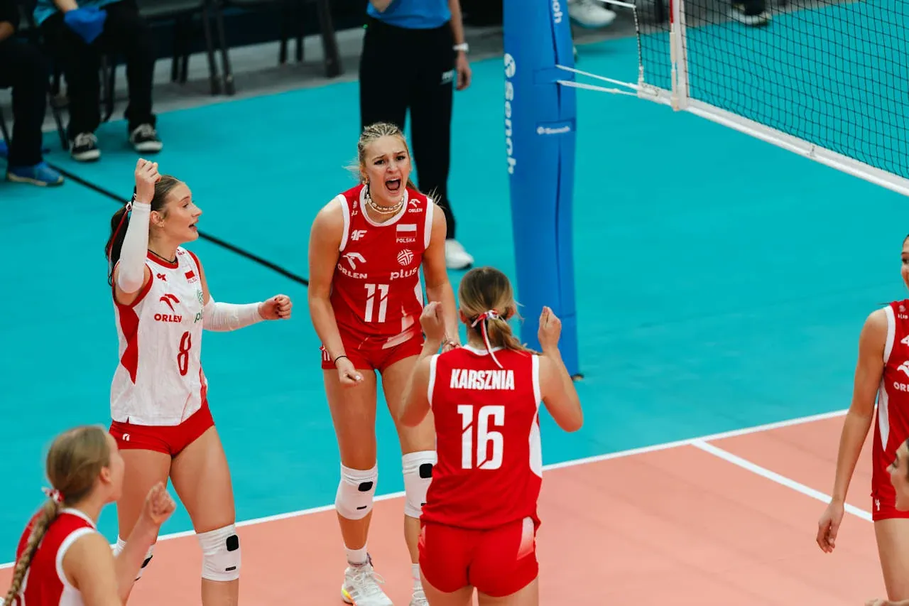 Zawodniczki drużyny siatkarskiej w czerwono-białych strojach świętujące punkt na boisku, otoczone siatką i liniami boiska.
