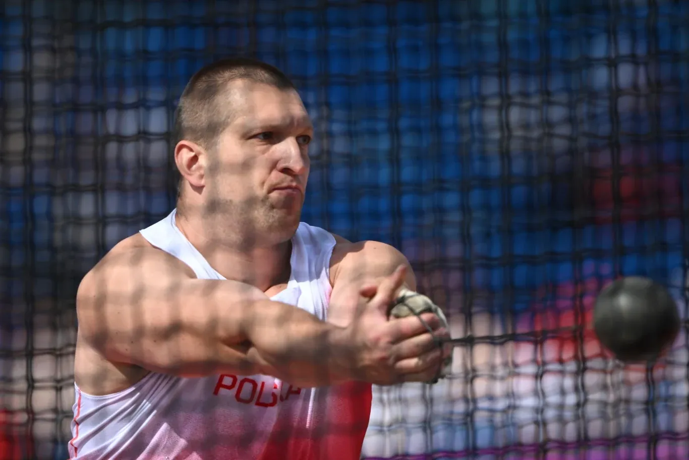 Zawodnik w białym stroju sportowym z napisem 'Polska' w trakcie rzutu młotem, z wyrazem skupienia na twarzy, podczas zawodów lekkoatletycznych.