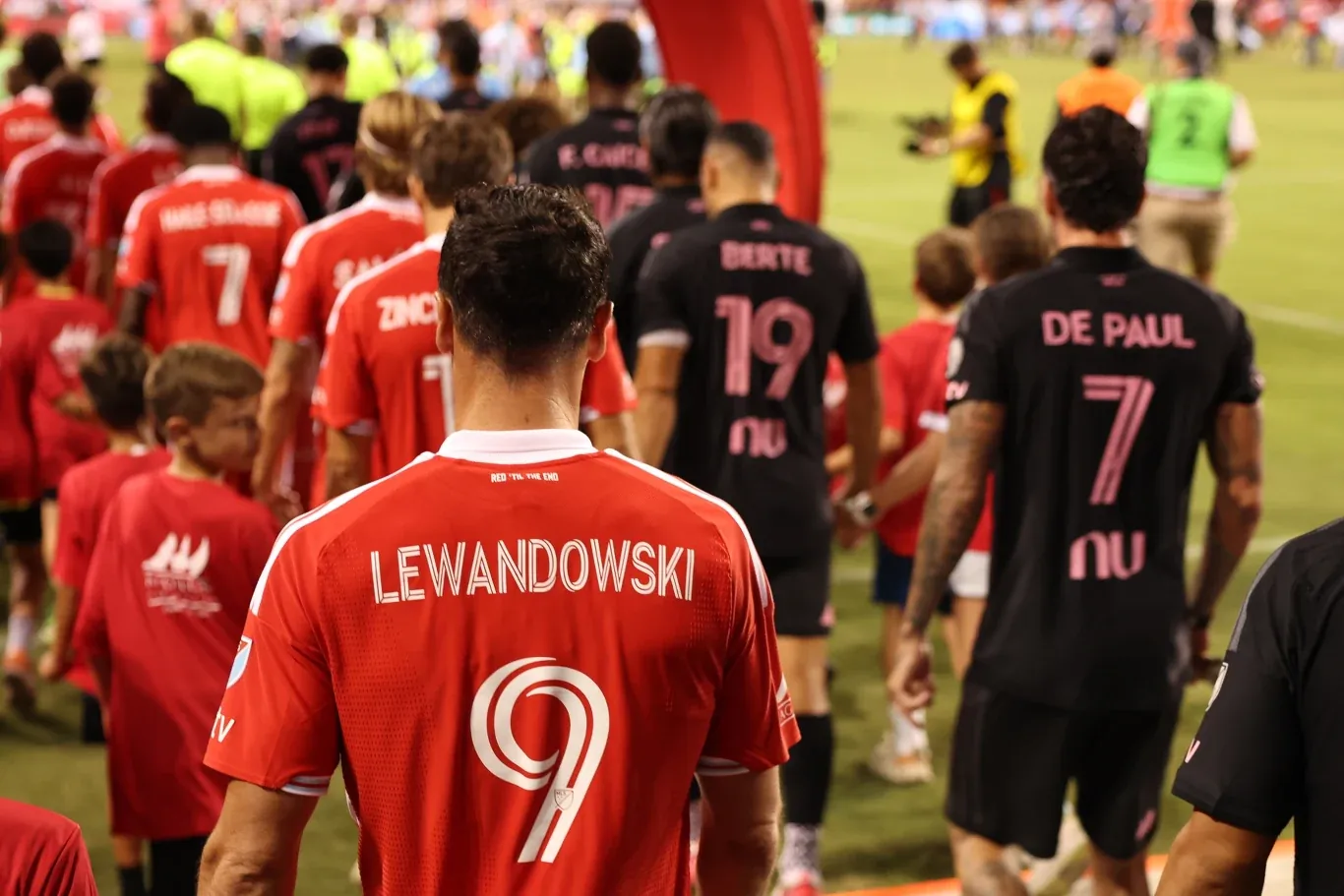 Piłkarz w czerwonej koszulce z nazwiskiem Lewandowski i numerem 9 idzie na boisko wśród innych zawodników ubranych w stroje różniące się barwami, pośród dzieci i obsługi sportowej.