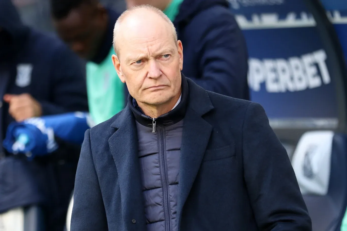 Łysy mężczyzna w granatowym płaszczu i pikowanej kurtce stoi na tle stadionu piłkarskiego, z poważnym wyrazem twarzy, w tle rozmyte postacie i fragmenty reklamy.